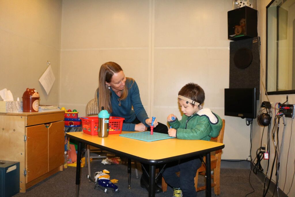 Audiology assistant supporting a child with hearing loss in an audiology booth during a testing session.