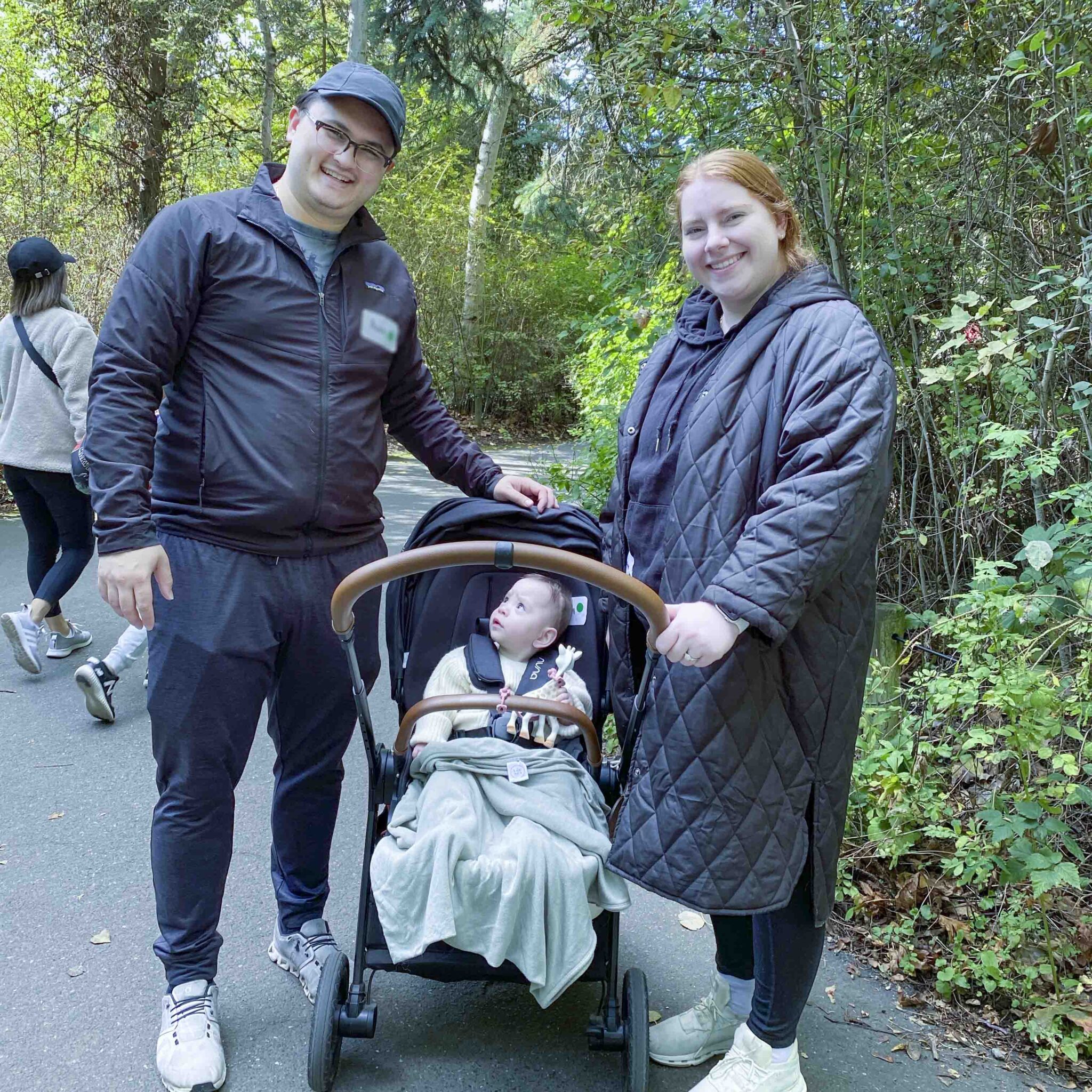 Family group photo of two adults and one child sitting in a stroller looking up at their father as they're walking on a paved pathway in the woods at the zoo.