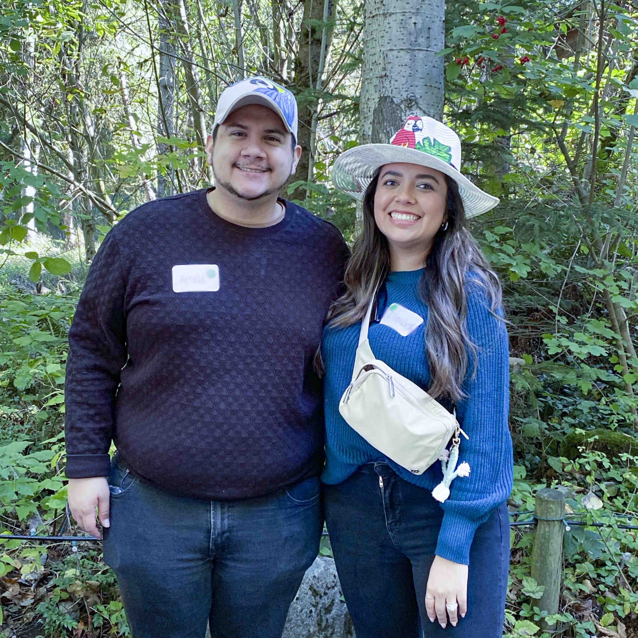 Couple smile for camera as they stand in front of a wooded forest
