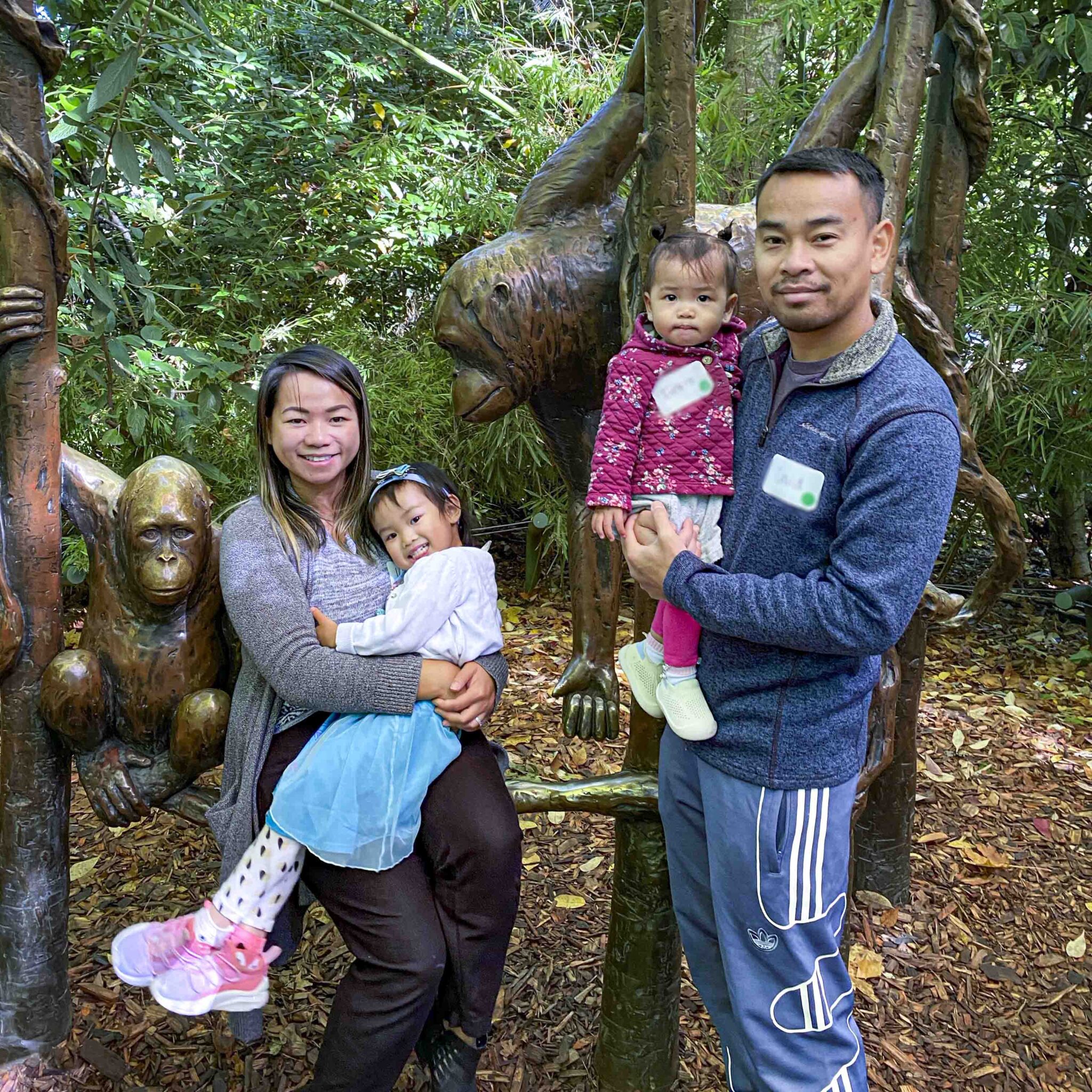 family group photo of two adults and two children standing/sitting in front of a sculpture inside a wooded zoo exhibit.